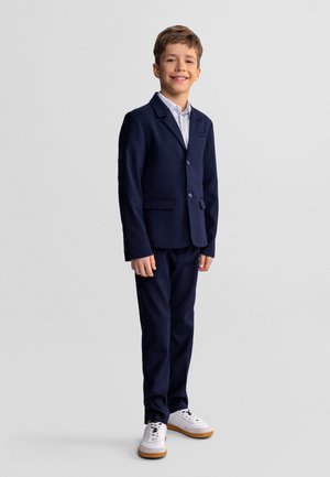 Uśmiechnięty chłopiec stojący, ubrany w granatowy garnitur z białą koszulą i białymi trampkami na jednolitym jasnym tle.