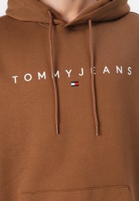Bruine katoenen hoodie met witte tekst "TOMMYJEANS" en een klein rood, wit en blauw logo. Voorzien van een trekkoord en een voorzak.