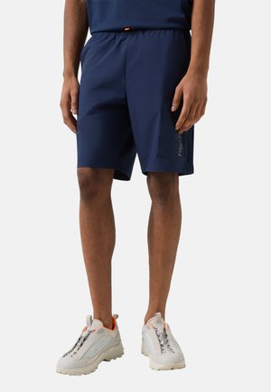 Man wearing navy blue cargo shorts with side pocket and white sneakers, standing against plain background.