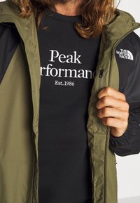 Chaqueta verde oliva con mangas negras, bolsillos con cremallera y el logo de The North Face. Debajo lleva un sudadera negra con texto blanco.