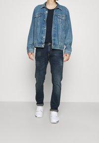 Světle modrá džínová bunda se dvěma předními kapsami. Kombinováno s tmavým tričkem a tmavými džíny. Bílí tenisky dokončují outfit.