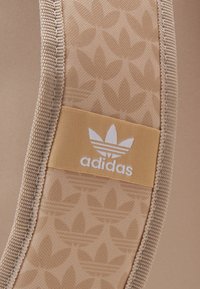 Béžový textilní panel s jemným listovým vzorem a obdélníkovou žlutou nálepkou s bílým logem "adidas" a detaily se třemi pruhy.