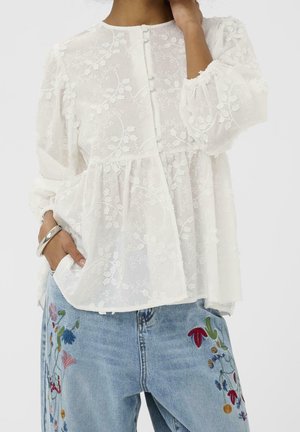 Donna che indossa una camicetta bianca trasparente con ricami floreali e bottoni in tessuto abbinati, abbinata a jeans azzurri con ricami floreali colorati.