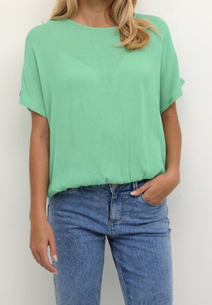 Femme portant un chemisier ample à manches courtes vert menthe, rentré dans un jean bleu taille haute, une main dans la poche.