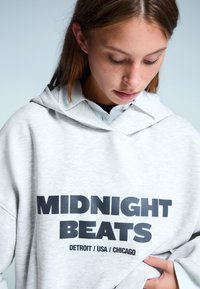 Svetle šedá mikina s kapucňou s veľkým čiernym nápisom "MIDNIGHT BEATS" spolu s "DETROIT / USA / CHICAGO" na prednej strane. Mäkká textúra, uvoľnený strih.
