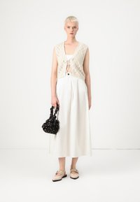 Beige knitted vest with open back and tie closure, white high-waisted skirt, black patterned bag, and light-colored flat shoes.