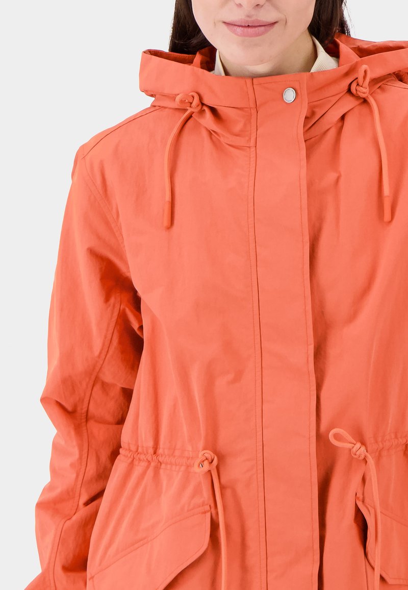 Vrouw in een feloranje hooded jacket met verstelbare touwtjes en grote klepzakken, die lichtjes glimlacht tegen een effen achtergrond.