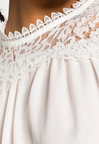 Ljusbeige topp med intrikata vita spetsdetaljer vid halsringningen och plisserad nederdel. Spetsen ger textur och detalj.