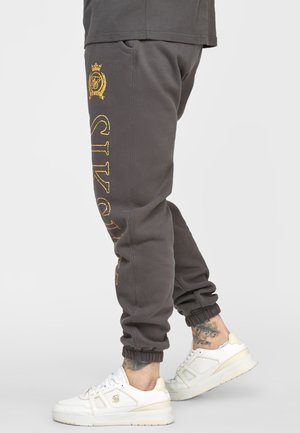 Person trägt dunkelgraue Jogginghose mit gelbem vertikalem Text an einem Bein und weiße Sneakers, sichtbare Tattoos an den Knöcheln.