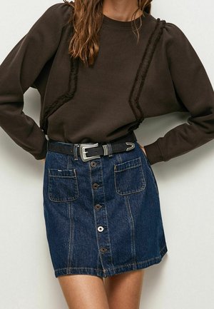 Femme portant un haut à manches longues marron foncé avec des rayures diagonales texturées, rentrée dans une jupe en jean à boutons sur le devant avec poches appliquées et ceinture noire.