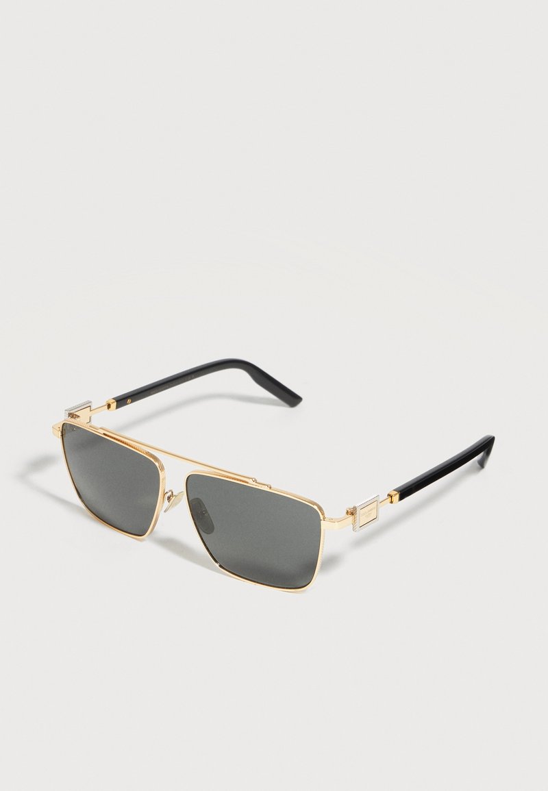 Ορθογώνια γυαλιά ηλίου με χρυσό πλαίσιο, σκοτεινούς φακούς και μαύρα άκρα στα σκέλη, ακουμπισμένα σε μία φωτεινή επιφάνεια.