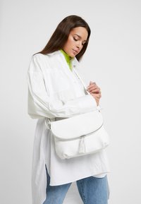 Weiße Lederhandtasche mit runder Form, Reißverschluss und Quasten-Detail. Quer über den Körper getragen, kombiniert mit einem übergroßen weißen Hemd und blauen Jeans.