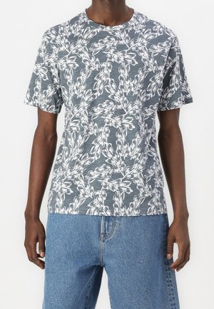 Personne portant un t-shirt gris à manches courtes avec un motif de branches feuillues blanches sur l'ensemble et un jean en denim bleu clair.