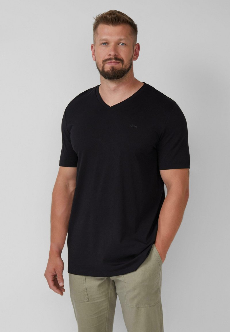 Baard man in een zwart T-shirt met korte mouwen en V-hals, en lichtbeige broek, staande met één hand in zijn zak tegen een eenvoudige achtergrond.
