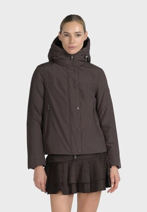 Femme portant une veste à capuche marron foncé avec des boutons sur le devant et des poches zippées, associée à une jupe noire superposée sur un fond uni.