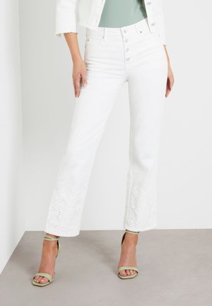 Kobieta w białych dżinsach z wysokim stanem z haftowanymi rozszerzanymi nogawkami i jasnobeżowozielonych sandałach na obcasie z paskiem wokół kostki.