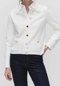 Bílé kardigan s límcem, se zlatými knoflíky a dvěma předními kapsami. Vyrobeno z hladké látky s utaženým střihem. V kombinaci s tmavými džíny.