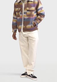 Veste multicolore à motifs avec un col pelucheux, associée à un pantalon crème et des baskets noires. Comprend des poches avant et une coupe décontractée.