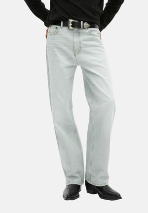 Persoon in lichtblauwe rechte jeans met een zwarte riem en zwarte enkellaarzen met hakken, met de handen op de heupen.