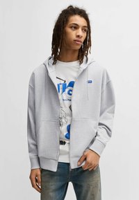 Gray zip-up hoodie made of cotton blend with a kangaroo pocket, ribbed cuffs, and a small blue logo. Worn over a white graphic t-shirt.