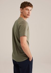 Khaki színű, rövid ujjú póló, puha, sima anyaggal és klasszikus szabással. A modell oldalról áll, kezei a zsebében, semleges háttér előtt.