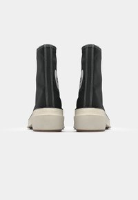 Juodi aukšti kanvės sportbačiai su balta siūle ir apvaliu logotipu kiekvienoje išorinėje pusėje, turintys smėlio spalvos guminius padus.