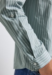 Close-up van een groene langermige shirt met witte verticale strepen en witte knopen, gedragen door een persoon met blauwe jeans.