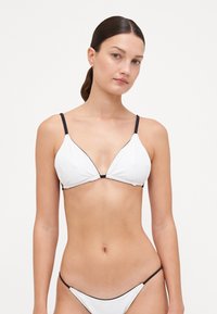 Hvit bikinitopp med trekantkopper, tynne svarte stropper og svart kant. Glatt tekstur, minimalistisk design og ingen synlig metall.