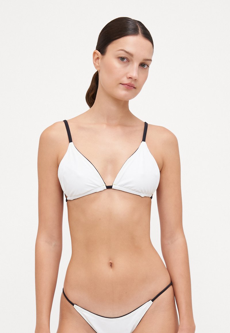 Hvit bikinitopp med trekantkopper, tynne svarte stropper og svart kant. Glatt tekstur, minimalistisk design og ingen synlig metall.