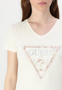 T-shirt crème en V en coton, avec un logo triangulaire en rose doré métallisé orné de strass et le texte "GUESS U.S.A." en argent.
