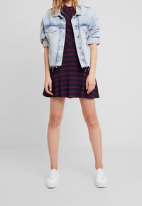Giubbotto di jeans con maniche arrotolate su un vestito a trapezio a righe blu navy e rosse. Sneakers bianche completano il look. Design semplice, vestibilità casual.
