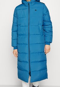 Lang blå dunfrakke med hætte, quiltet design, frontlynlås og sidelommer. Har elastiske manchetter og et logo på brystet.