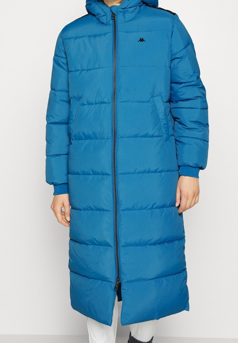 Lang blå dunfrakke med hætte, quiltet design, frontlynlås og sidelommer. Har elastiske manchetter og et logo på brystet.