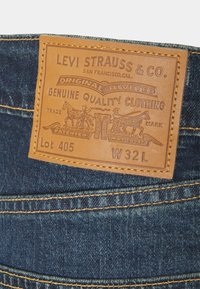 Étiquette en cuir Levi Strauss & Co. sur un jean en denim bleu indiquant "Original Riveted", taille W32 L, lot 405, avec deux chevaux tirant sur un jean.
