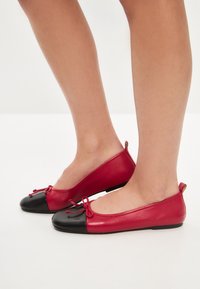 Ballerine in pelle rossa con punta nera, dotate di un fiocco decorativo, texture liscia e suola bassa e piatta.