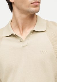 Homme portant un polo en maille beige avec le logo "BOSS" sur la poitrine, légèrement tourné de côté, sur un fond blanc uni.