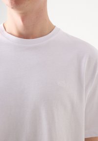 T-shirt en coton blanc avec un col rond et des manches courtes. Logo brodé discret sur la poitrine. Texture lisse.