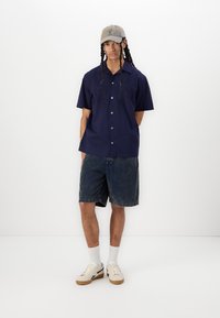 Camisa de manga corta azul marino con detalles bordados, combinada con pantalones cortos de mezclilla. El modelo lleva una gorra beis y zapatillas blancas con toques en tonocafé.