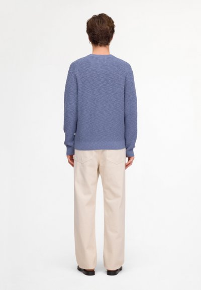 Homme portant un pull en maille bleu, un pantalon large crème et des chaussures noires, debout de dos devant un fond blanc uni.