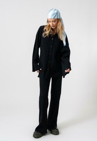 Zwarte geribbelde gebreide oversized knoopshirt gecombineerd met flared zwarte broek. Model draagt een blauw-wit gestreepte sjaal. Chunky zwarte platform schoenen.