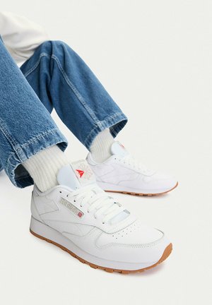 Osoba ubrana w białe trampki Reebok, białe skarpetki i niebieskie dżinsy, siedząca z nogami zgiętymi na tle jednolitej białej ściany.