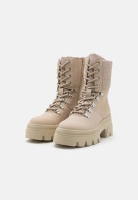 Botas de tobillo beige con parte superior texturizada, diseño con cordones y cuello acolchado. Presentan una suela gruesa con surcos profundos para mayor tracción.