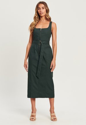 Dark green sleeveless dress with a square neckline, front zipper, and a waist tie featuring a circular buckle. Smooth texture; knee-length.
