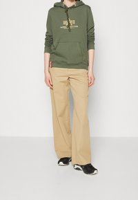 Felpa verde oliva con scritte dorate, tasca a marsupio, pantaloni beige a gamba larga e sneakers nere con accenti bianchi. Vestibilità semplice e rilassata.