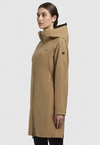 Beige lange jas met een capuchon, voorzien van een voorrits, zijzakken en een zwart logo accent op de mouw. Glad, waterafstotend materiaal.