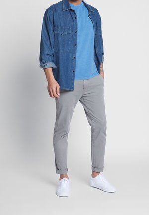 En denim skjorte med knapper, en blå t-shirt, lysegrå bukser og hvide sneakers. Skjorten har to frontlommer. Bukserne er rullet op ved anklerne.