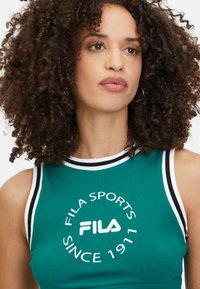 Mujer con cabello rizado que lleva una camiseta sin mangas de deporte FILA verde con ribetes en blanco y negro, mirando ligeramente hacia un lado.