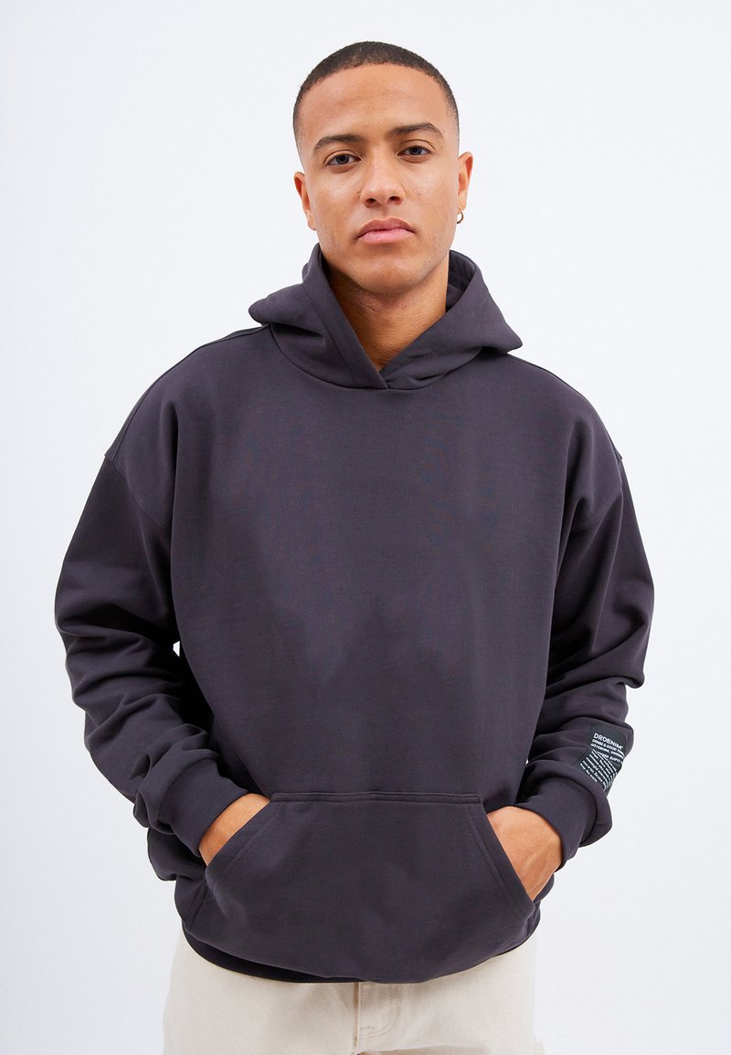 Ciemnoszara bluza z kapturem o luźnym kroju, z przednimi kieszeniami i małą metką na lewym rękawie. Miękka faktura materiału.