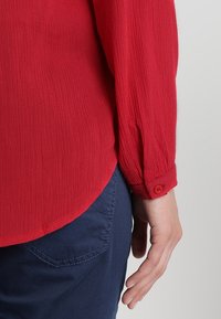 Camicia rossa a texture con polsini a bottone, orlo curvo e vestibilità rilassata, abbinata a pantaloni blu scuro. Focus principale sui dettagli del tessuto della camicia.