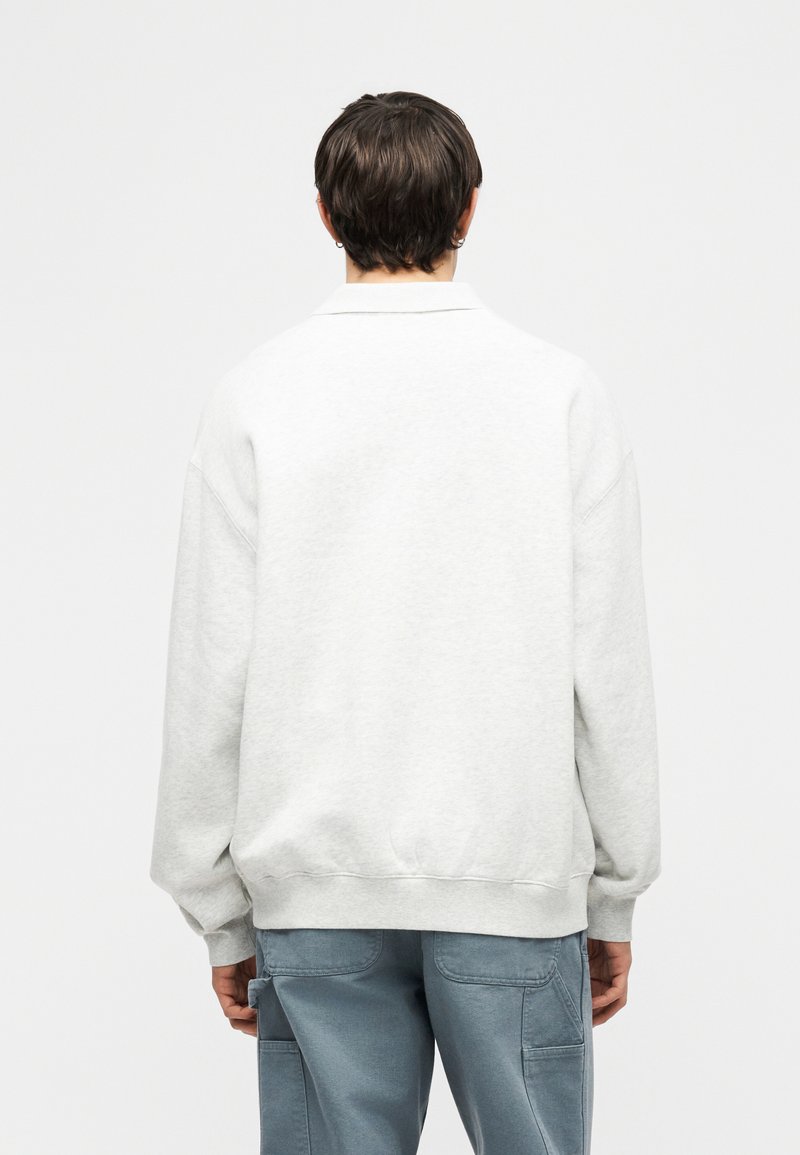 Person mit kurzen dunklen Haaren, die einen weißen Langarmpullover und blaue Cargohosen trägt, steht mit dem Rücken zu einem einfarbigen Hintergrund.